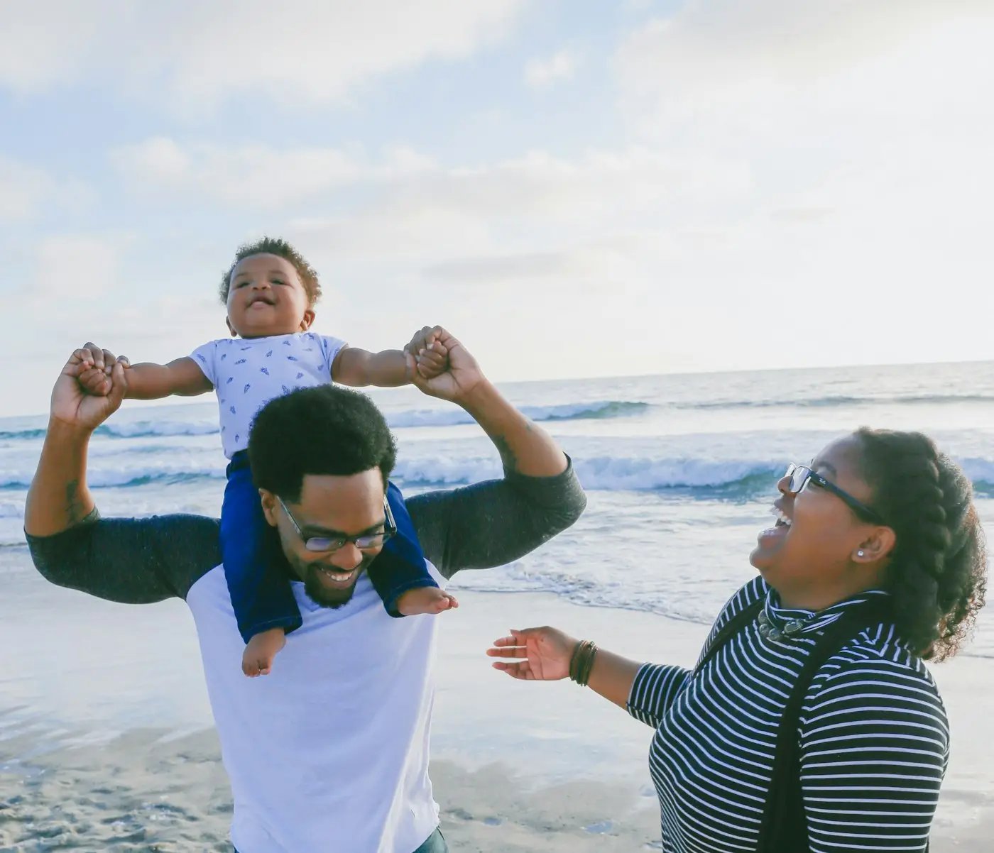 A father carries his laughing toddler on his shoulders while the mother laughs beside them at the beach.
