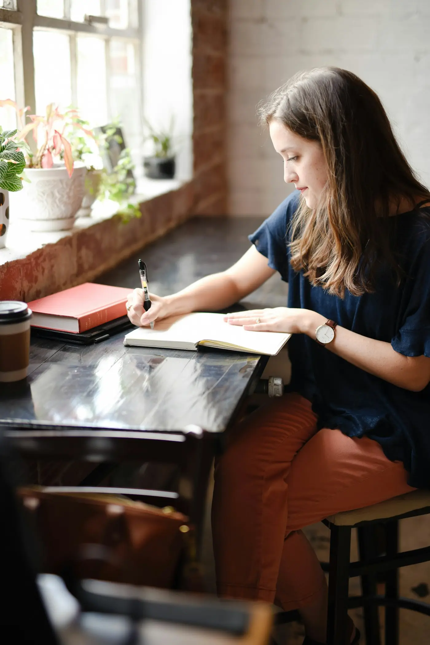 A woman sits by a sunlit window, writing in a journal with a cup of coffee and plants nearby.